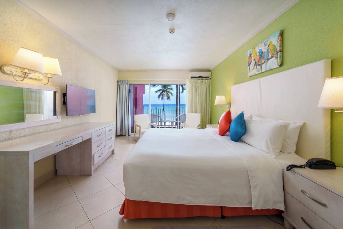 A hotel room with a bed, bedside tables, a desk, a chair, and a balcony with a view of the ocean.