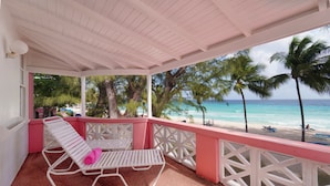 Een overdekt balkon met witte reling en twee loungestoelen, met uitzicht op een strand met palmbomen en de oceaan.