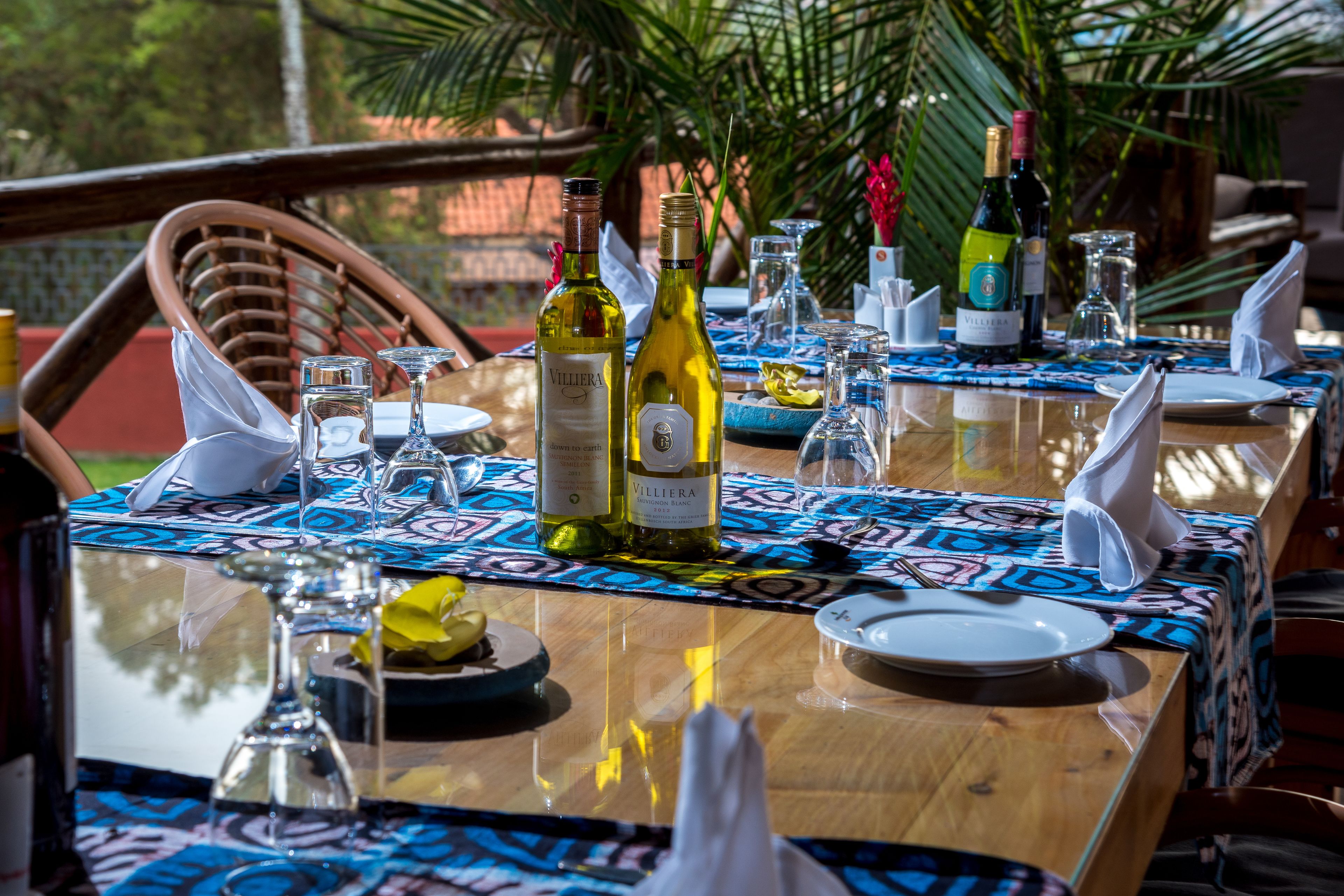 A dining table set with wine bottles, glasses, and plates, surrounded by chairs and a patterned tablecloth.