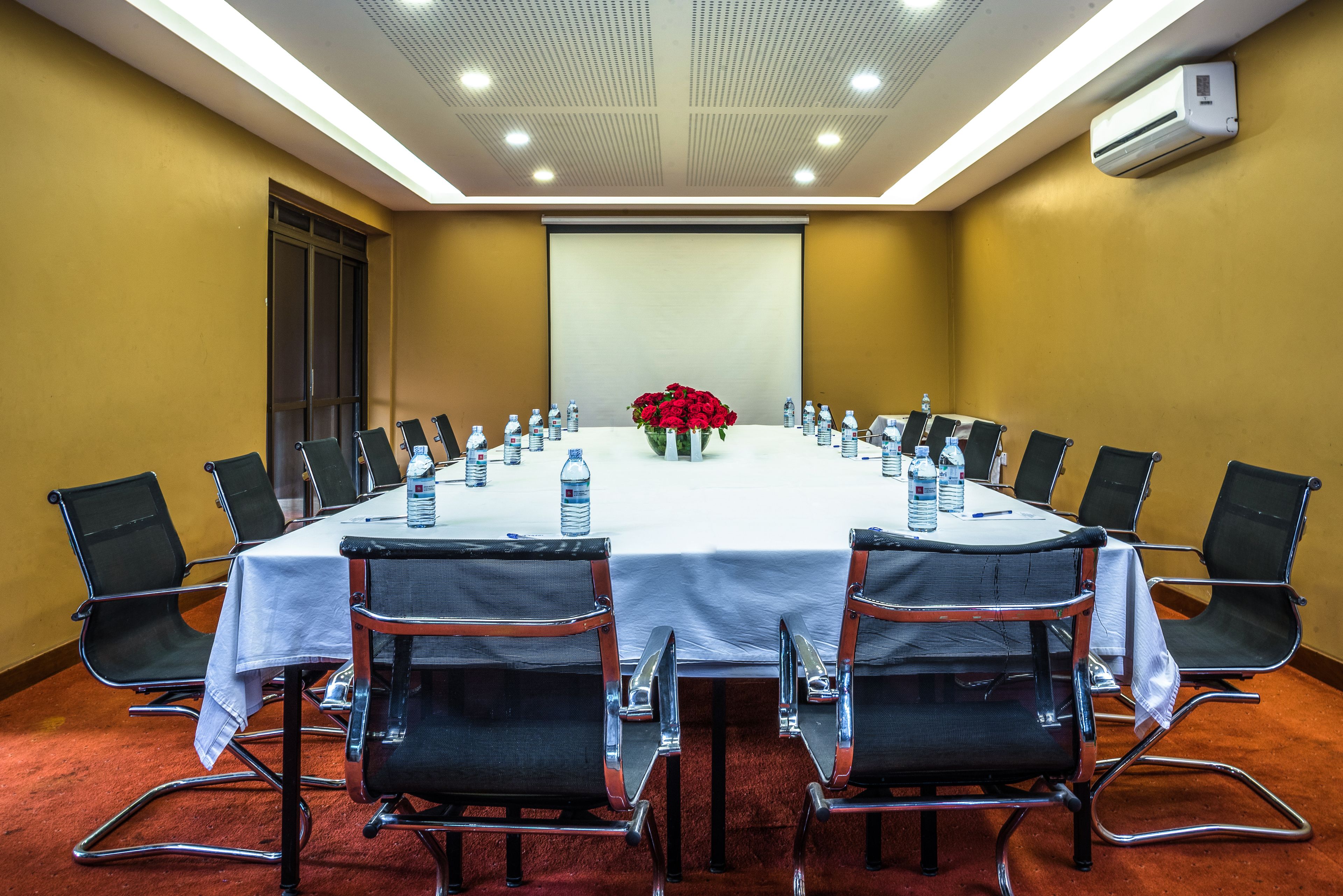 Un salón de conferencias con una larga mesa, sillas negras, botellas de agua y un centro de mesa floral.