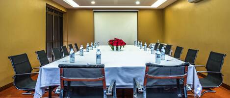 Un salón de conferencias con una larga mesa, sillas negras, botellas de agua y un centro de mesa floral.