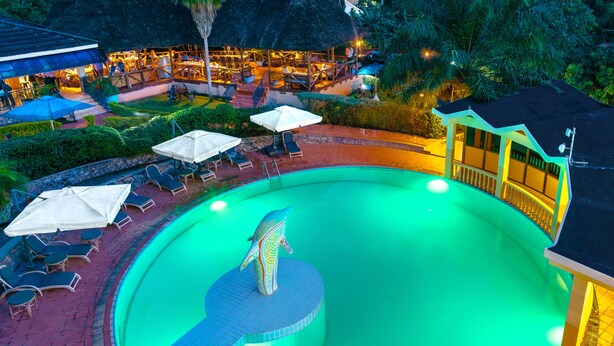 A swimming pool with lounge chairs and umbrellas, surrounded by a well-maintained garden and a building with a thatched roof. - Dolphin Suites (Kampala)