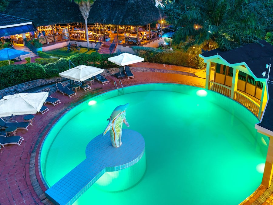 Ein Swimmingpool mit Liegestühlen und Sonnenschirmen, umgeben von einem gepflegten Garten und einem Gebäude mit einem Strohdach.