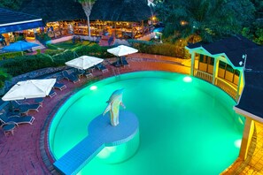 A swimming pool with lounge chairs and umbrellas, surrounded by a well-maintained garden and a building with a thatched roof.