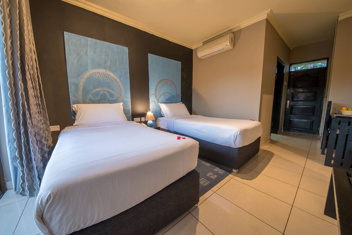 Une chambre d’hôtel avec deux lits, une grande fenêtre avec des rideaux, un climatiseur fixé au mur et une porte donnant sur un couloir.
