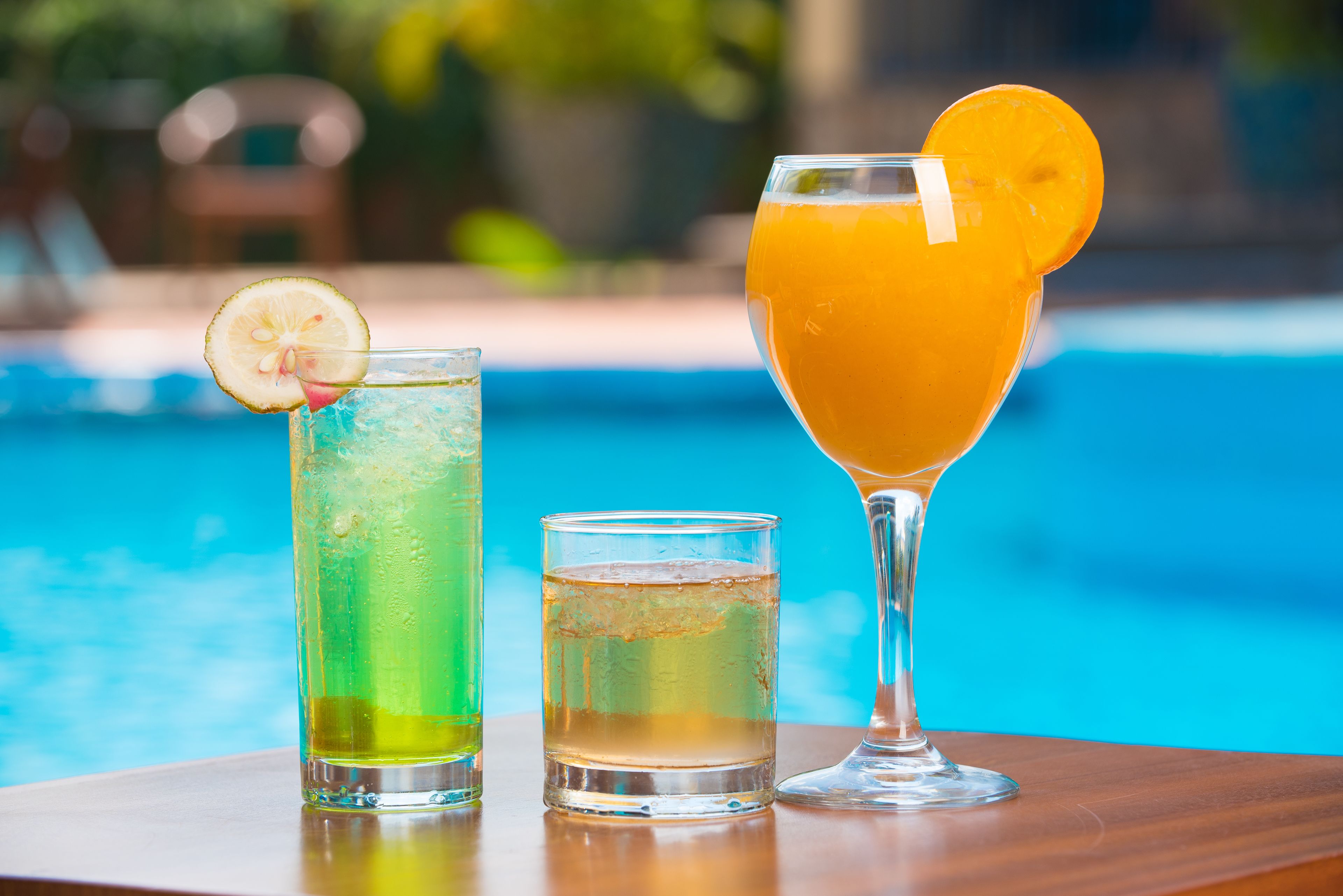 Three glasses of beverages on a wooden table by a pool.