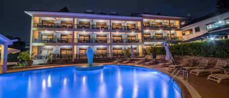 Un hotel de varios pisos con piscina, mobiliario exterior y una fachada bien iluminada.