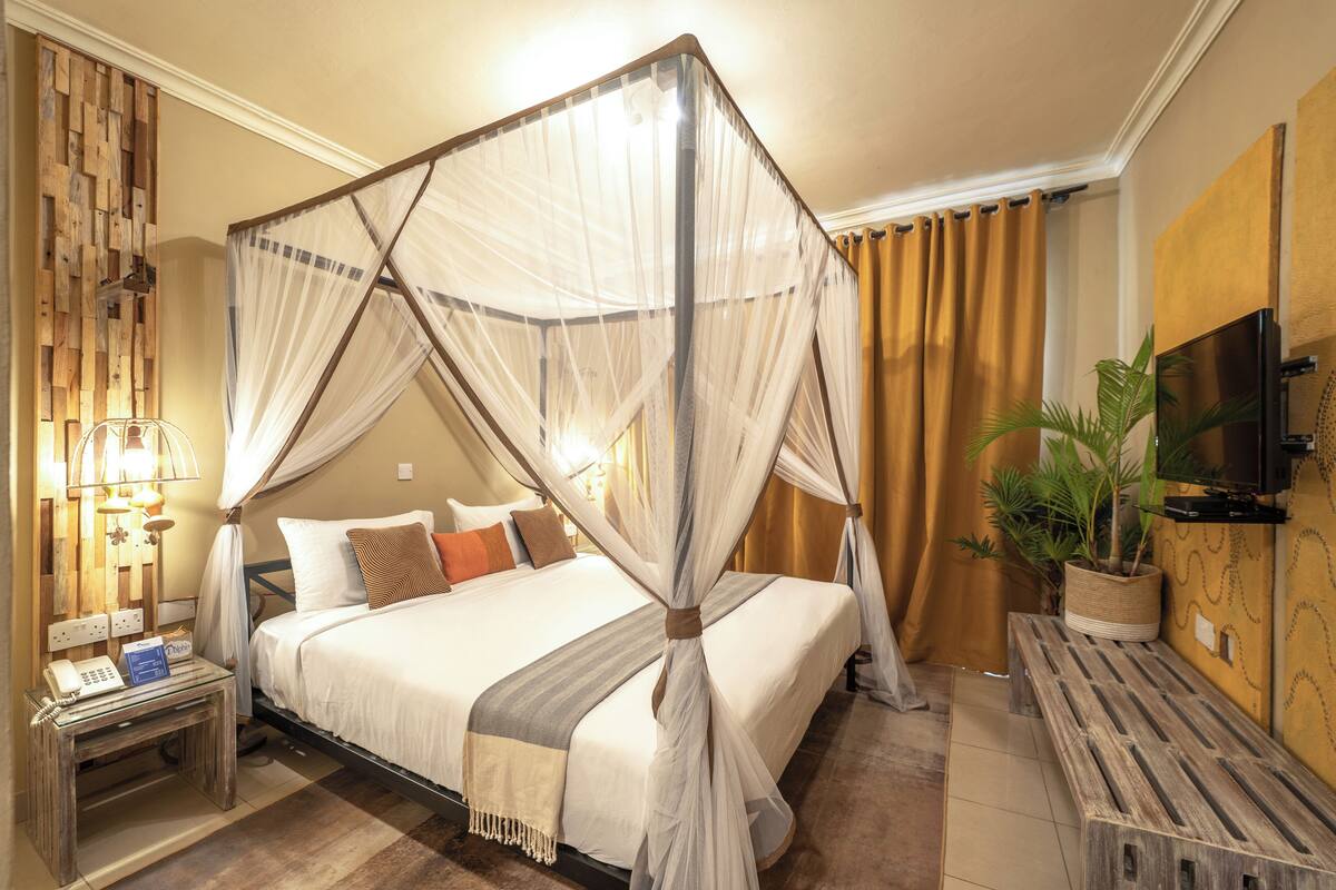 Une chambre d’hôtel avec un lit à baldaquin, une table de chevet avec une lampe, une télévision et une plante en pot.