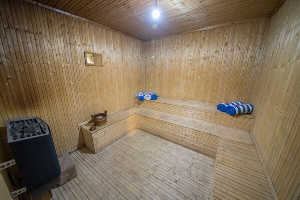 A wooden sauna room with benches, a stone heater, and a trash bin.