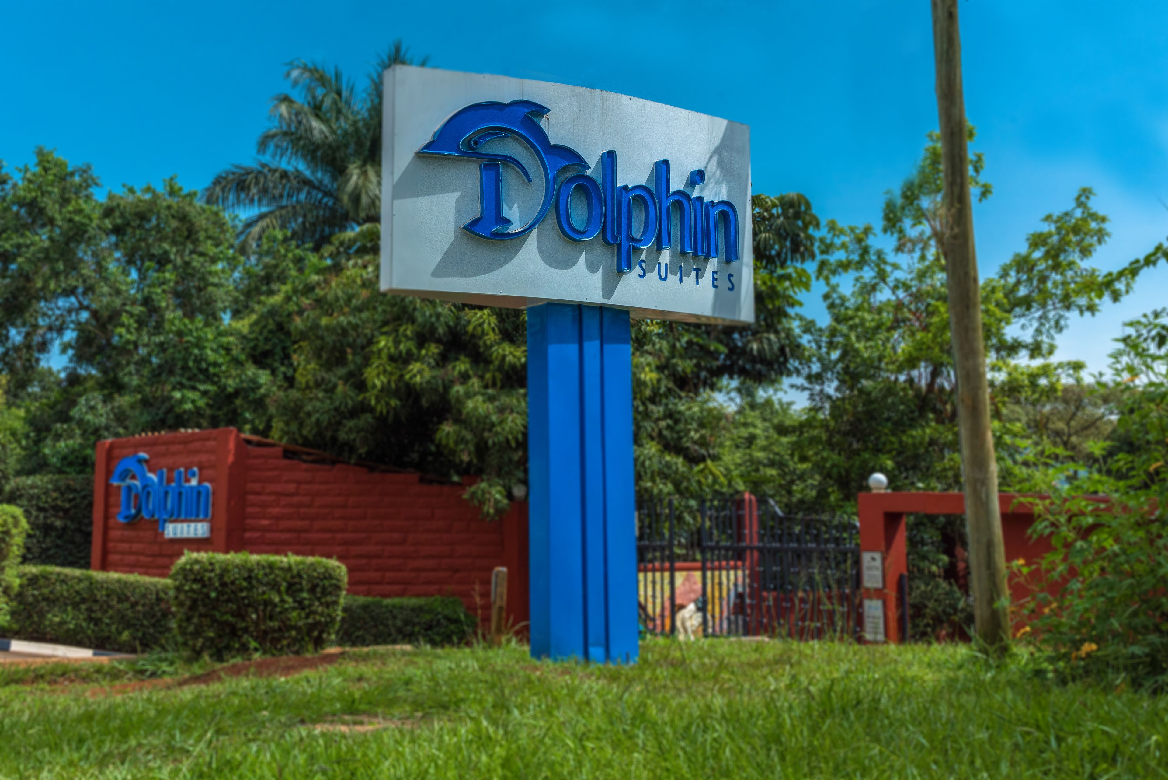A sign for Dolphin Suites with a blue and white color scheme, set against a backdrop of lush greenery and a clear blue sky.