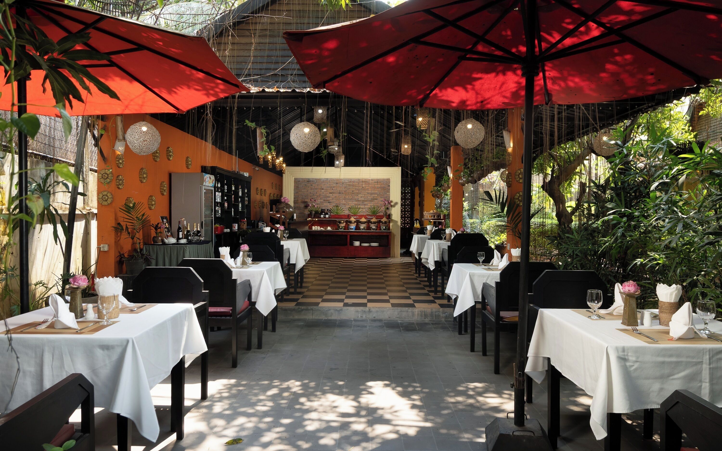 Un restaurante al aire libre con mesas y sillas, sombrillas rojas, luces colgantes y plantas en macetas.