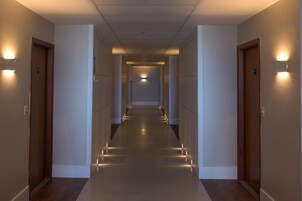 Um corredor comprido e estreito com portas em ambos os lados, iluminado por luzes fixadas na parede.