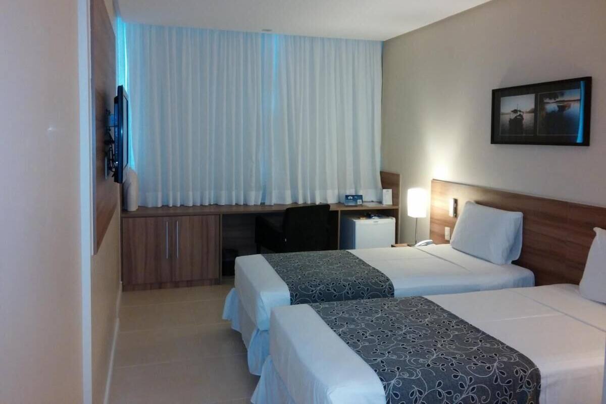 A hotel room with two beds, a desk, a TV, and a window with curtains.