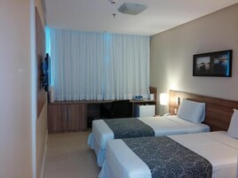 Quarto de hotel com duas camas, uma escrivaninha, televisão e janela com cortinas.
