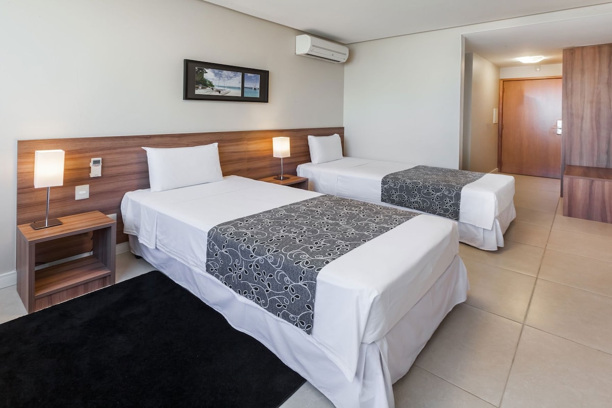 A hotel room with two beds, a nightstand with a lamp, a picture on the wall, and an air conditioning unit.