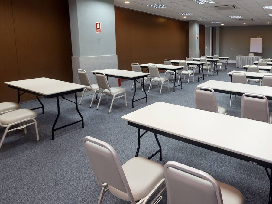 Een vergaderruimte met meerdere tafels en stoelen, een vloerbedekking en een whiteboard op de achtergrond.