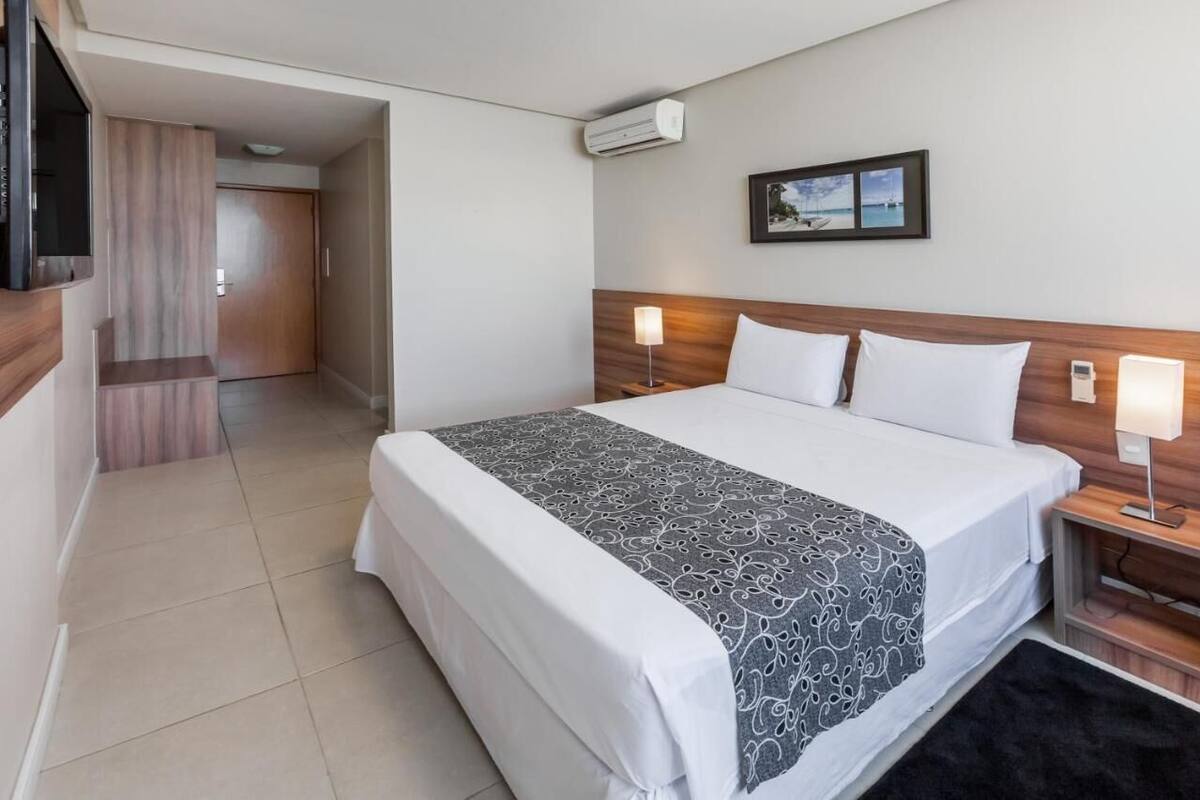 A hotel room with a large bed, two bedside tables with lamps, a wall-mounted air conditioner, and a framed picture above the bed.