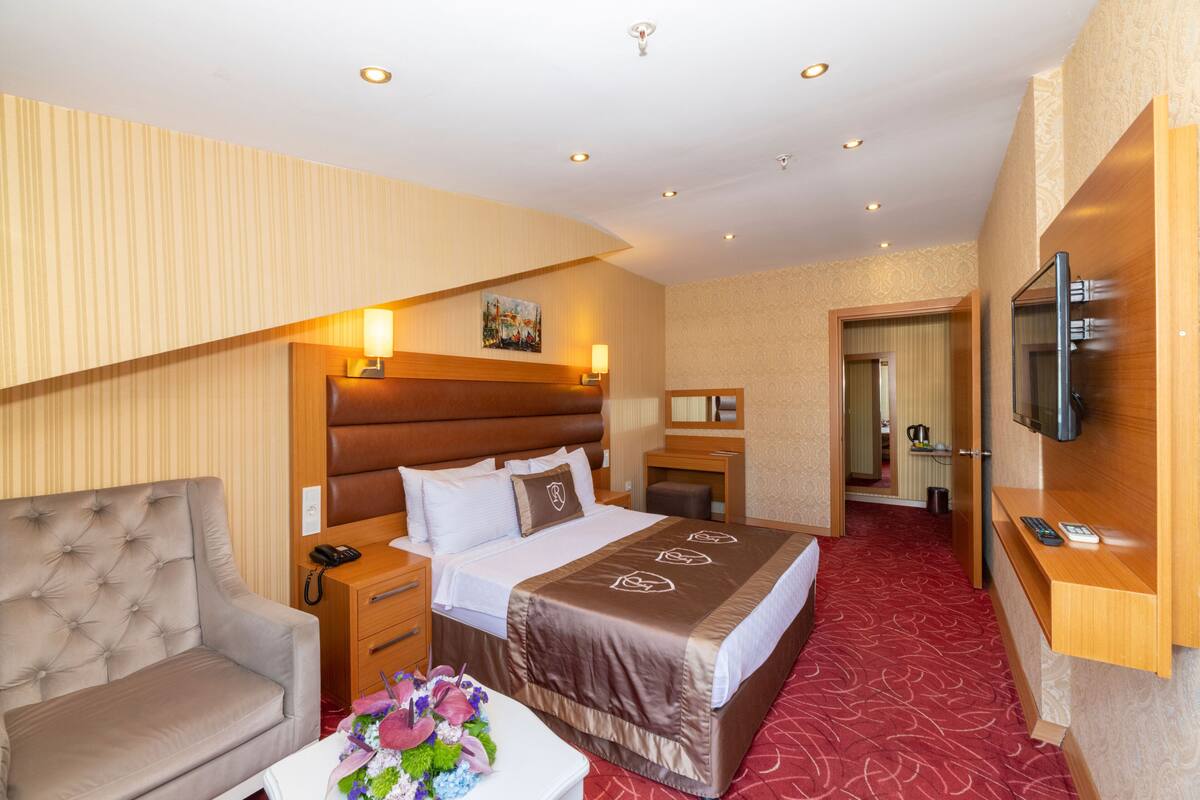 Ein Hotelzimmer mit einem großen Bett, einer Couch, einem Schreibtisch mit Lampe, einem Fernseher und einem durchgehenden Türspalt sichtbaren Badezimmer.