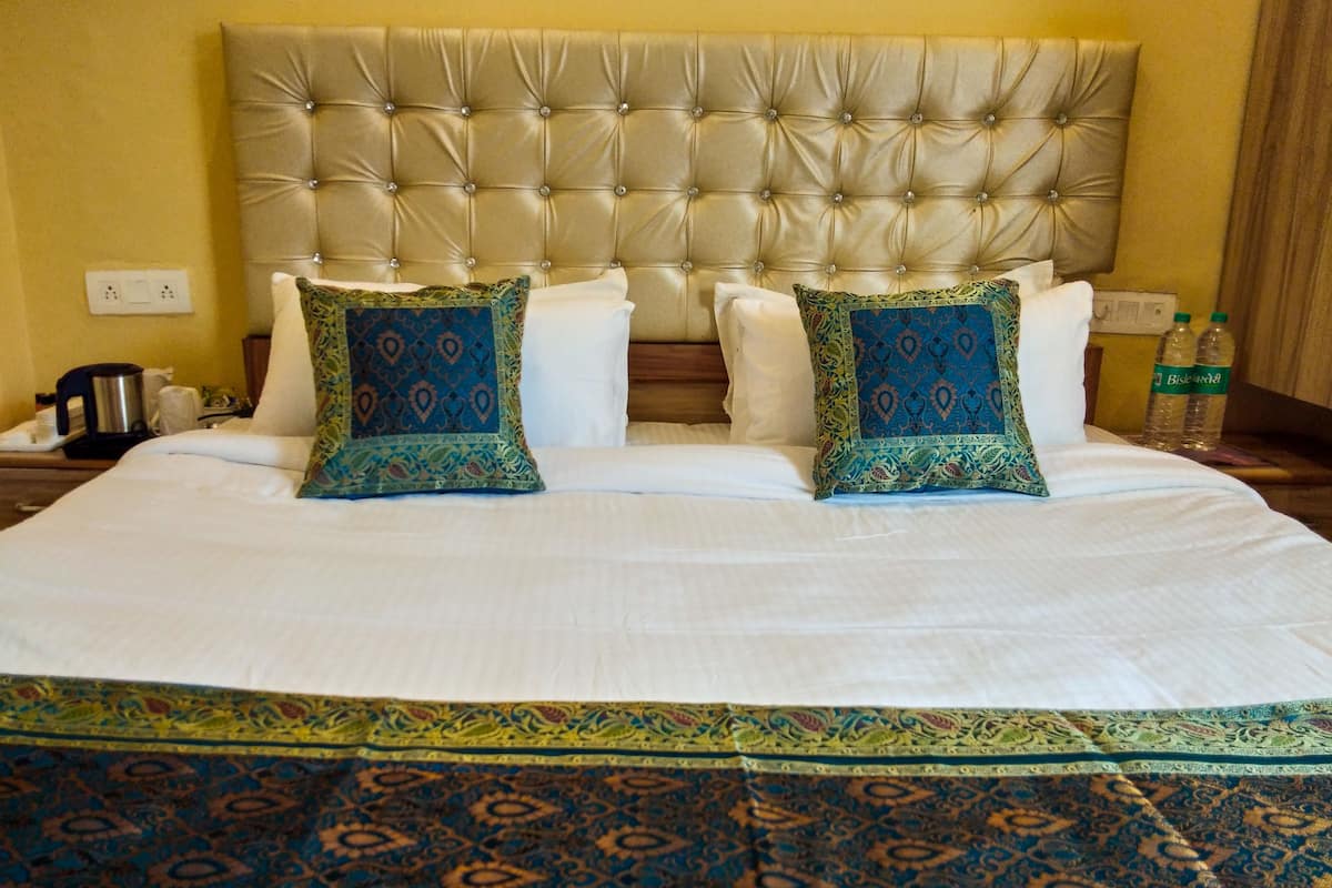 Habitación de hotel con una cama grande, dos almohadones decorativos, una mesita de noche con cafetera y una botella de agua.