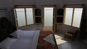 A room with a bed, a small table, and a bench. There are three windows providing a view of a body of water. - Palace on steps (Varanasi)
