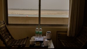 A room with a large window overlooking a beach. There are two wooden chairs, a small table with a tray holding two bottles of water, a white mug, and a white thermos. - Palace on steps (Varanasi)