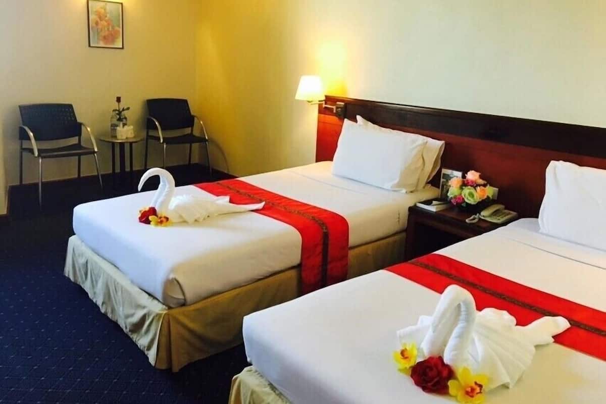 Zwei Hotelbetten mit weißen Laken, roten Bettdecken und schwanenförmigen Handtüchern. Im Zimmer befinden sich zwei Sessel und ein kleiner Tisch.