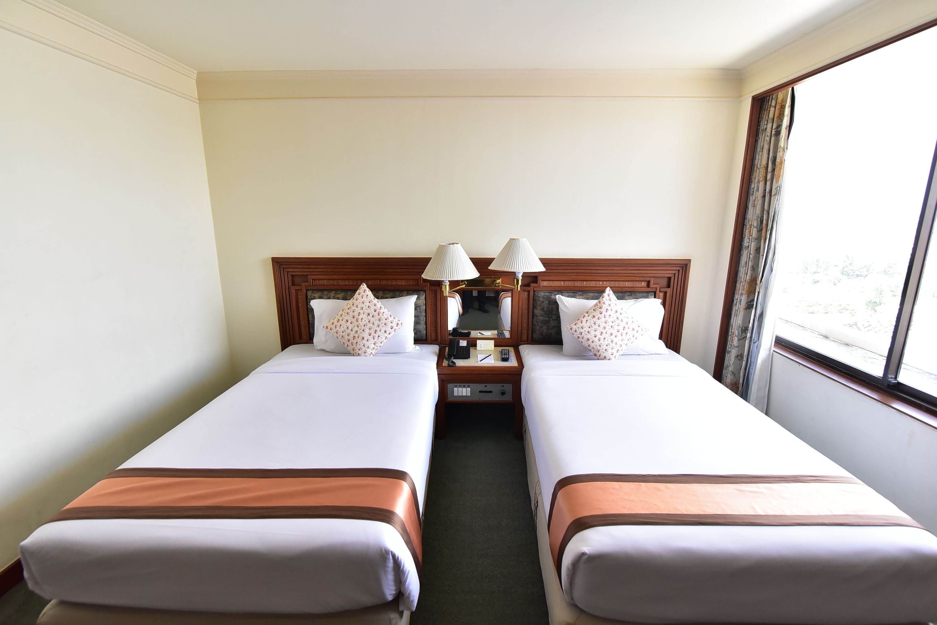 A hotel room with two single beds, a nightstand with lamps, a mirror, and a window with curtains.