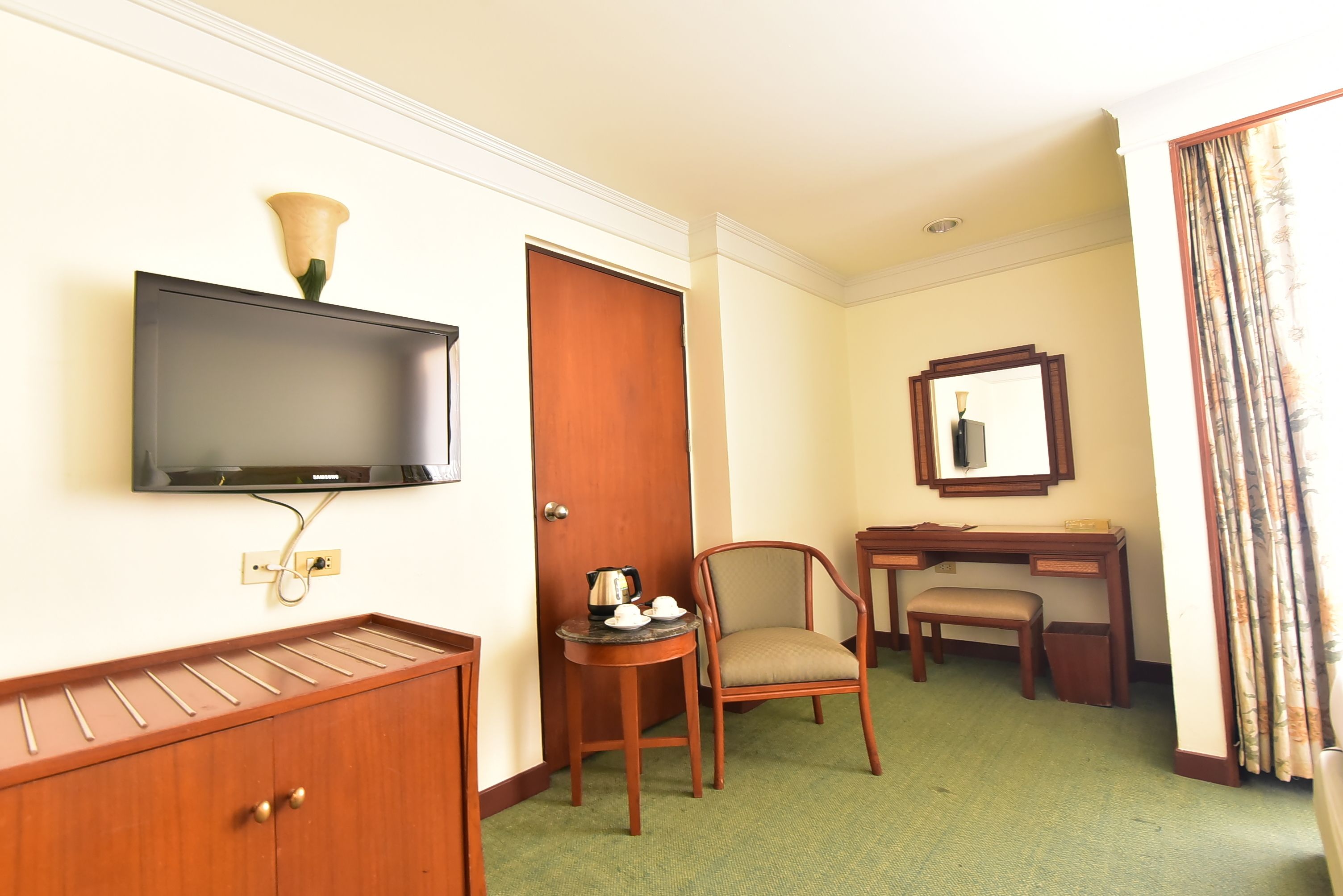 A hotel room with a flat-screen TV mounted on the wall, a wooden desk with a chair, a small table with a kettle and cups, and a green carpeted floor.
