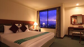 A hotel room with a large bed, a desk, a chair, and a view of the city at dusk. - Hansa JB Hotel (Hat Yai)