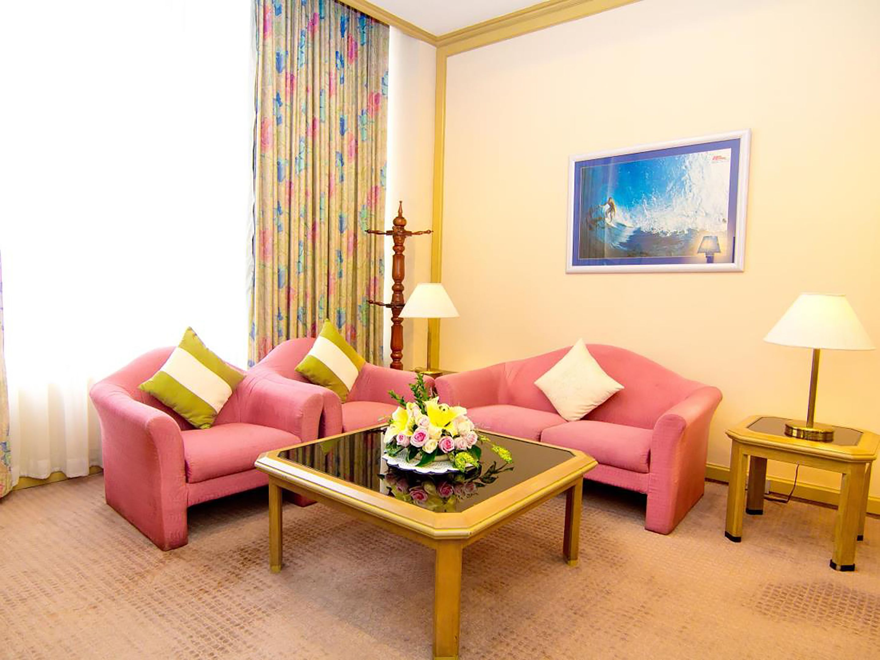 A room with two pink sofas, a glass coffee table with flowers, two lamps, a painting on the wall, and a curtain with a floral pattern.