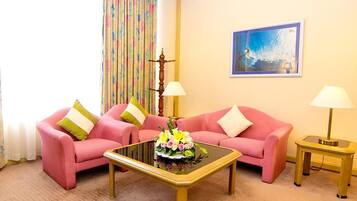 A room with two pink sofas, a glass coffee table with flowers, two lamps, a painting on the wall, and a curtain with a floral pattern.