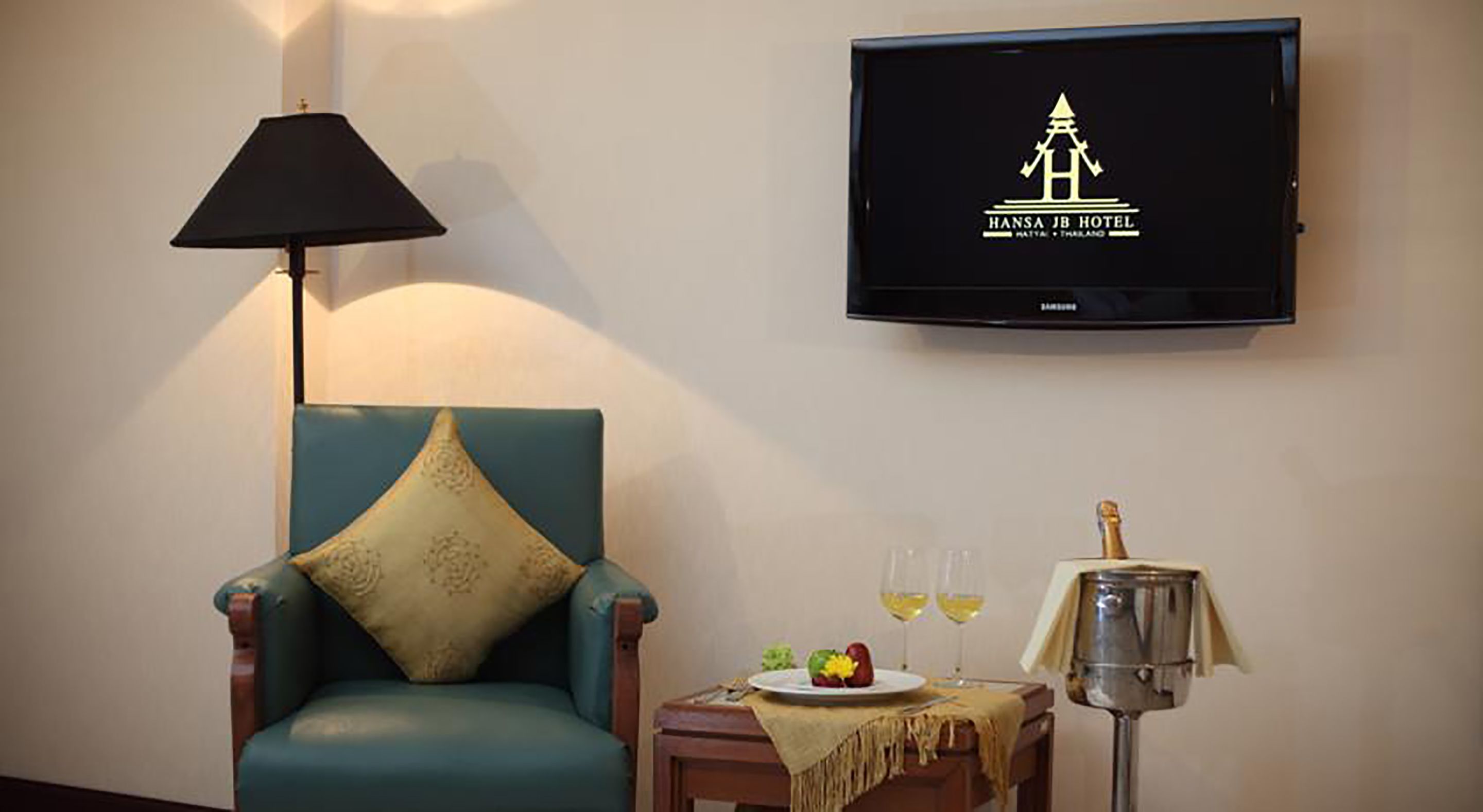 A teal armchair with a decorative pillow, a television displaying the Hansa JB Hotel logo, a small table with a plate of fruit and two glasses of wine, and a lamp.