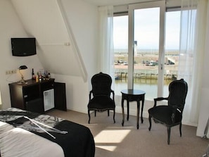 Ein Hotelzimmer mit einem Bett, einem Schreibtisch mit Lampe, zwei Stühlen, einem kleinen Tisch und einem gro ßen Fenster mit Blick auf den Strand.