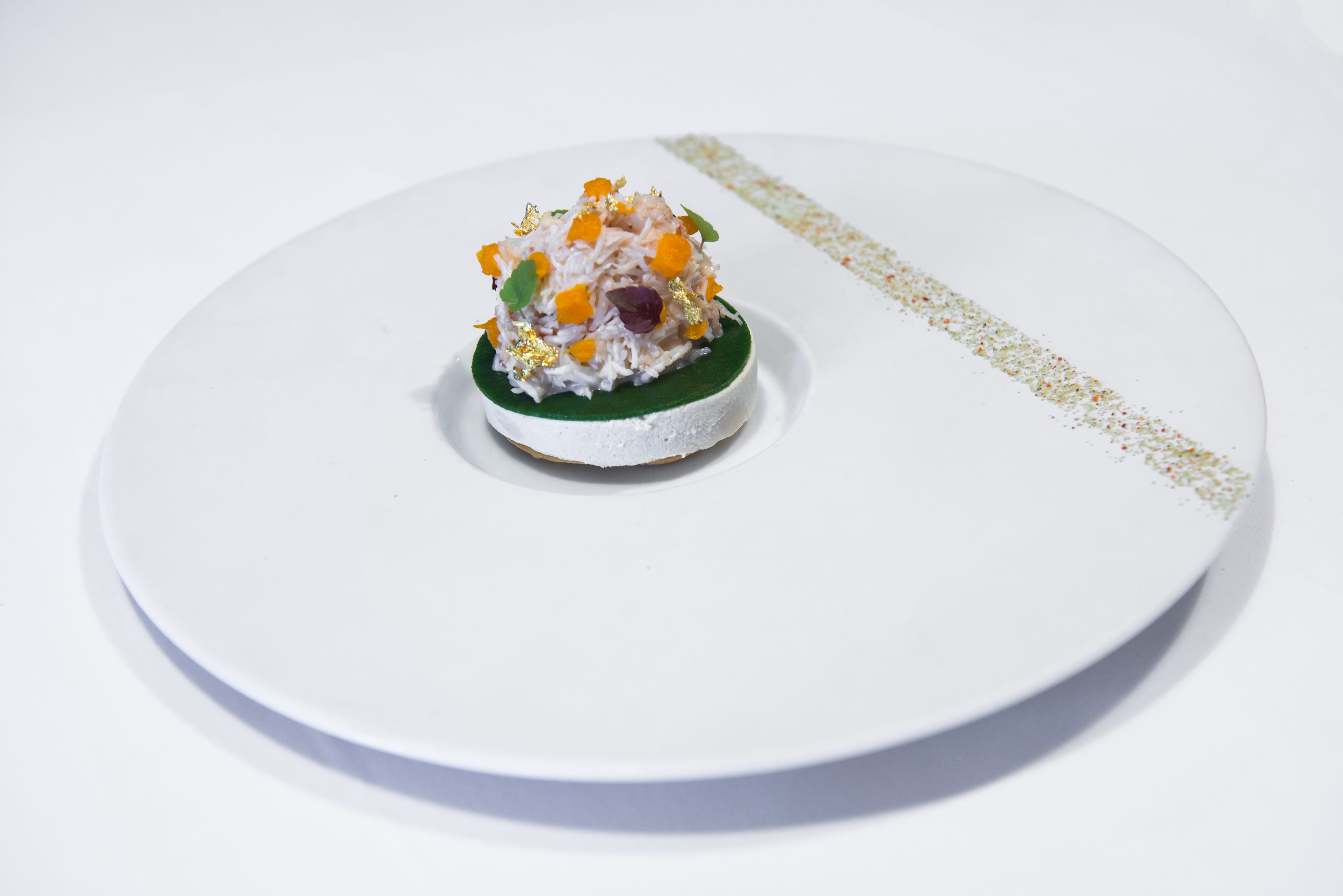 Een wit bord met een klein, rond, wit dessert op een groene ondergrond, versierd met goudvlokken en kleurrijke details.