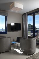 Una habitación moderna con un gran ventanal, un televisor de pantalla plana, mobiliario de mimbre y un ordenador portátil sobre una mesita.