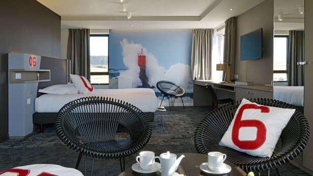 Habitación de hotel con cama, dos sillas, escritorio, televisor y un mural de un faro.
