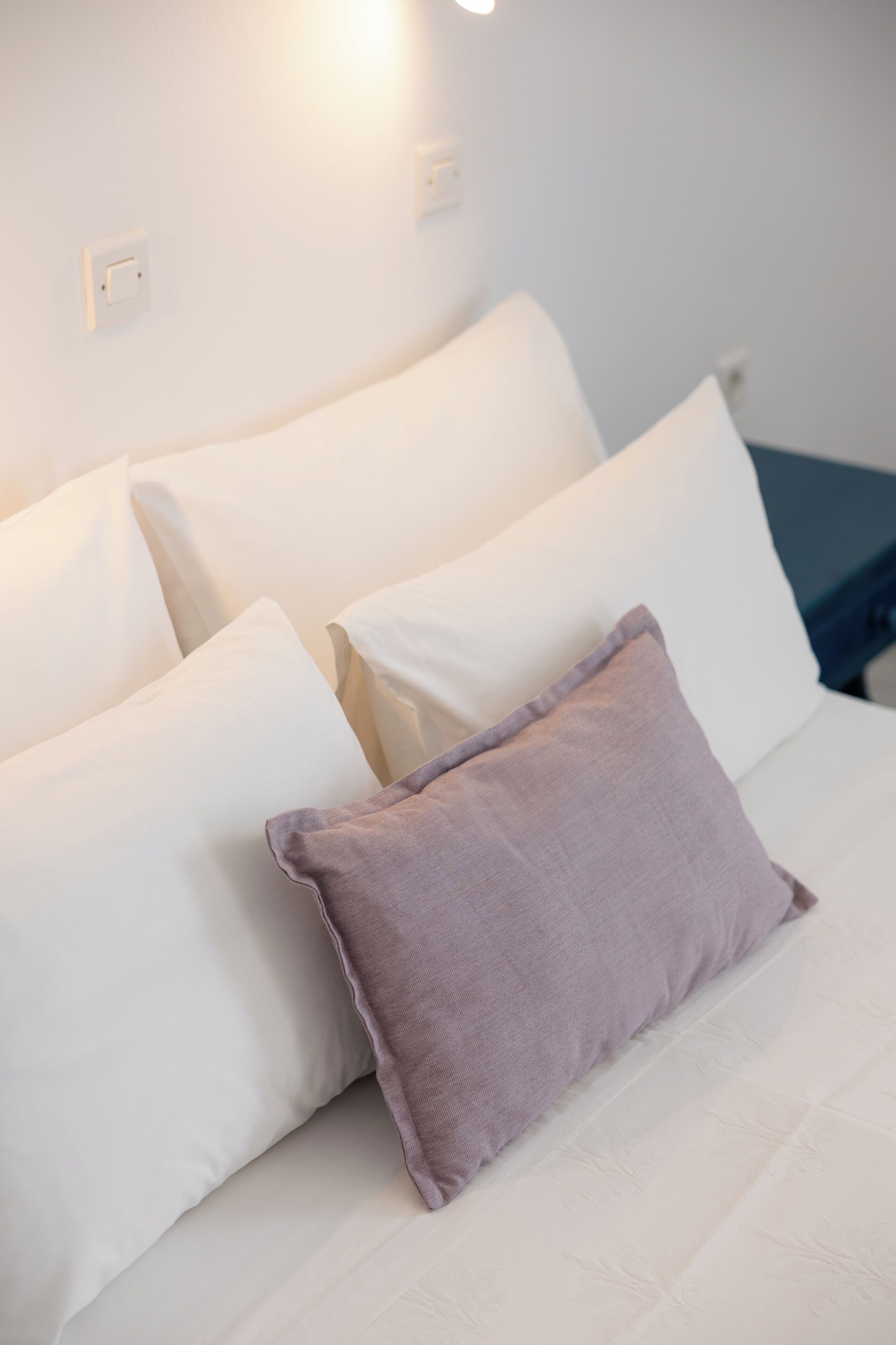 A bed with white linens and a single purple pillow.