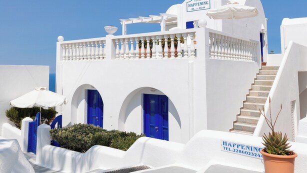 A white building with blue doors and a sign that reads "Villa Happening." - Villa Happening (Santorini)