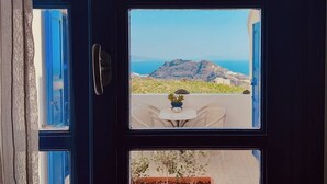 A dark wooden door with three windows. The top window shows a clear blue sky and a distant mountain range. The middle window reveals a patio with a table, chairs, and a potted plant. The bottom window displays a large orange planter with small plants. - Villa Happening (Santorini)