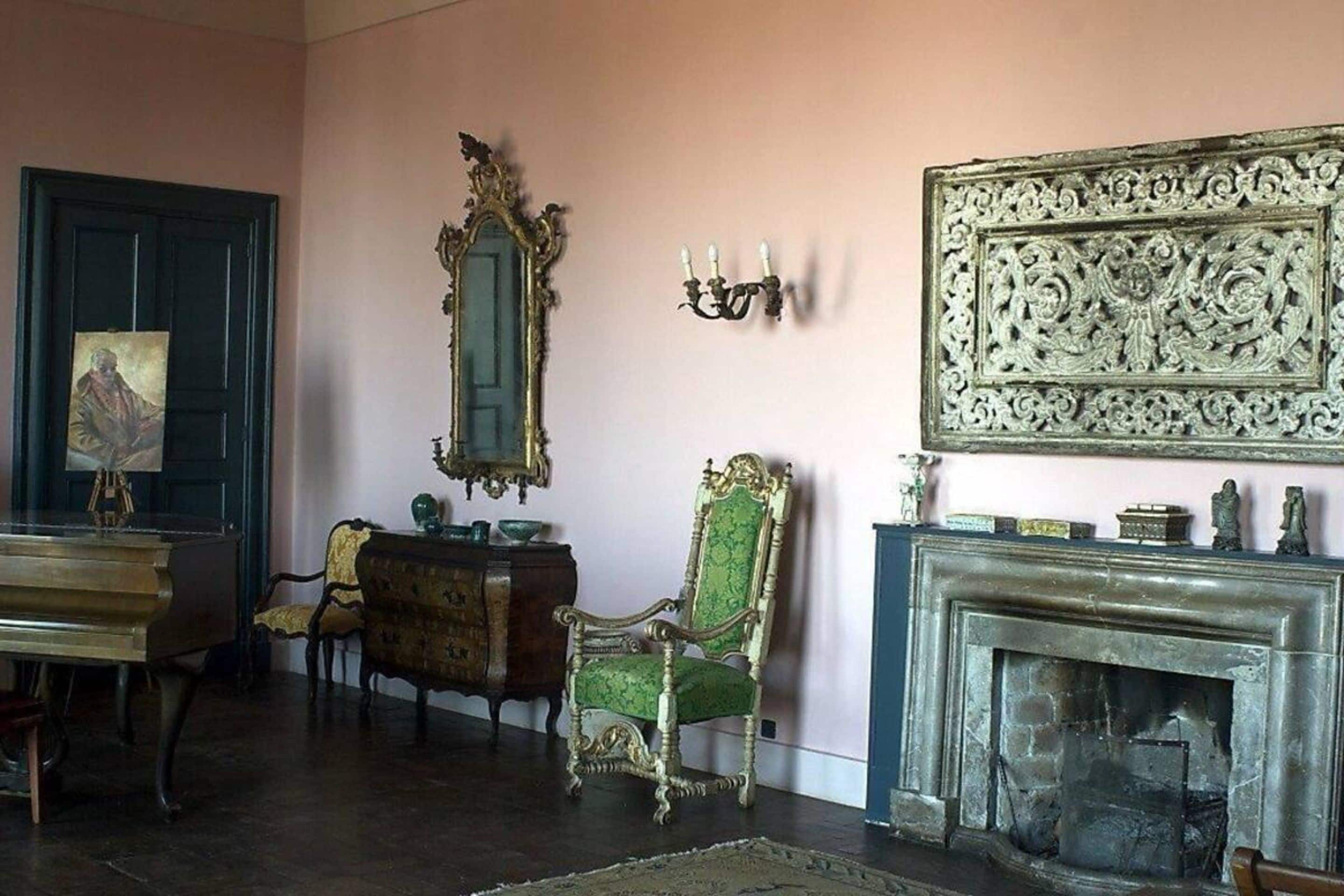 A room with a piano, a green upholstered chair, a mirror, a fireplace, and a wall-mounted candelabra.
