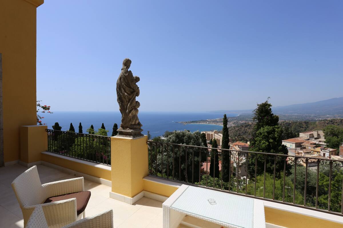 A balcony with wicker furniture, a statue, and a view of the sea and town.