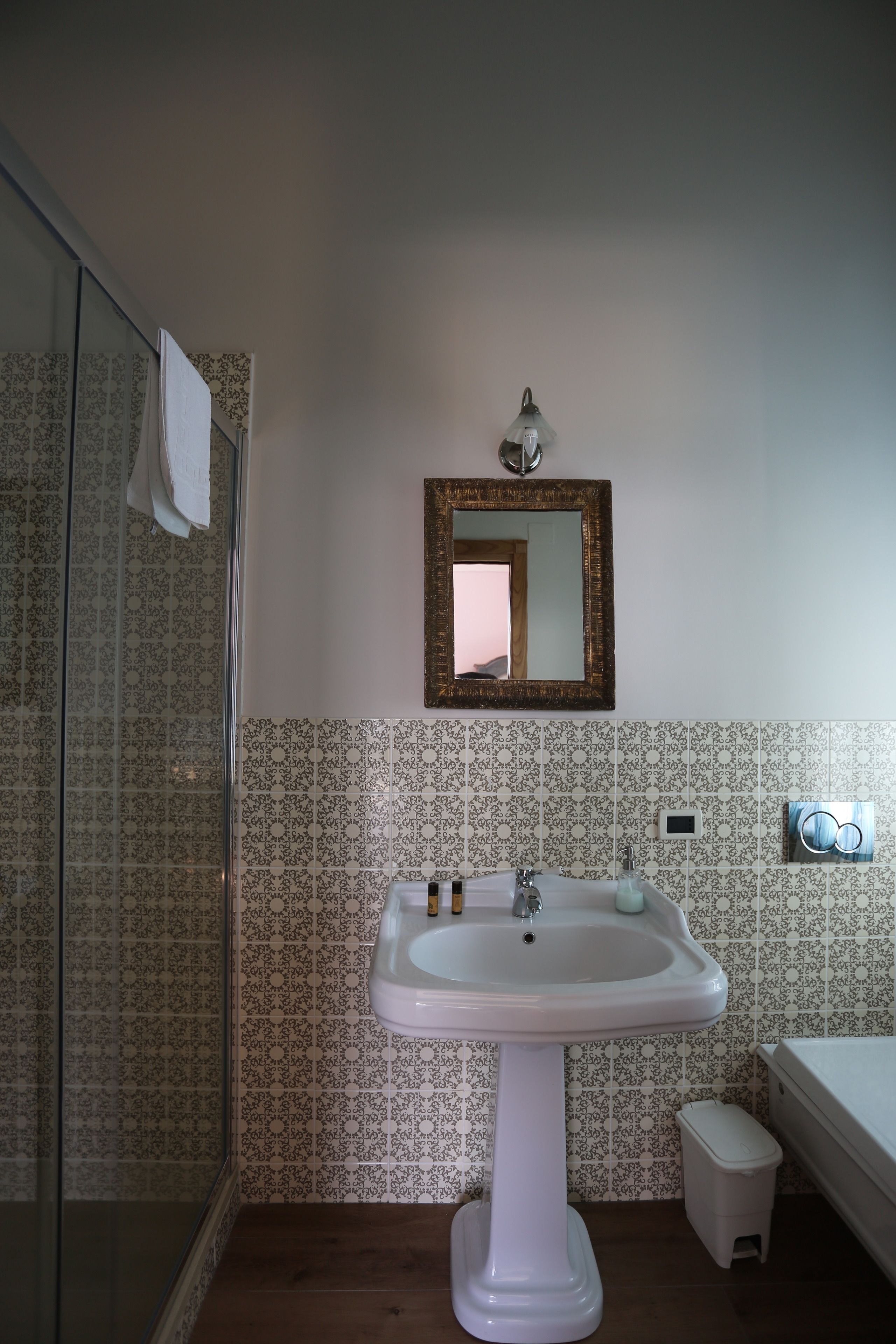 A bathroom with a pedestal sink, a glass shower enclosure, patterned tiles, a small trash bin, and a mirror with a decorative frame.
