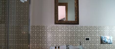 A bathroom with a pedestal sink, a glass shower enclosure, patterned tiles, a small trash bin, and a mirror with a decorative frame.