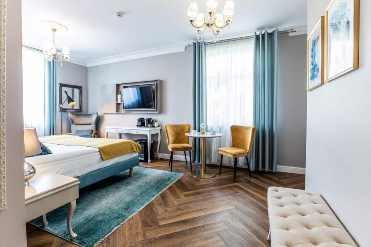A hotel room with a bed, a desk, two chairs, a TV, and a chandelier.