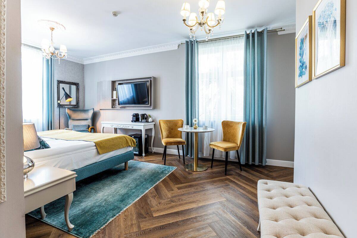 A hotel room with a bed, a desk, two chairs, a TV, and a chandelier.