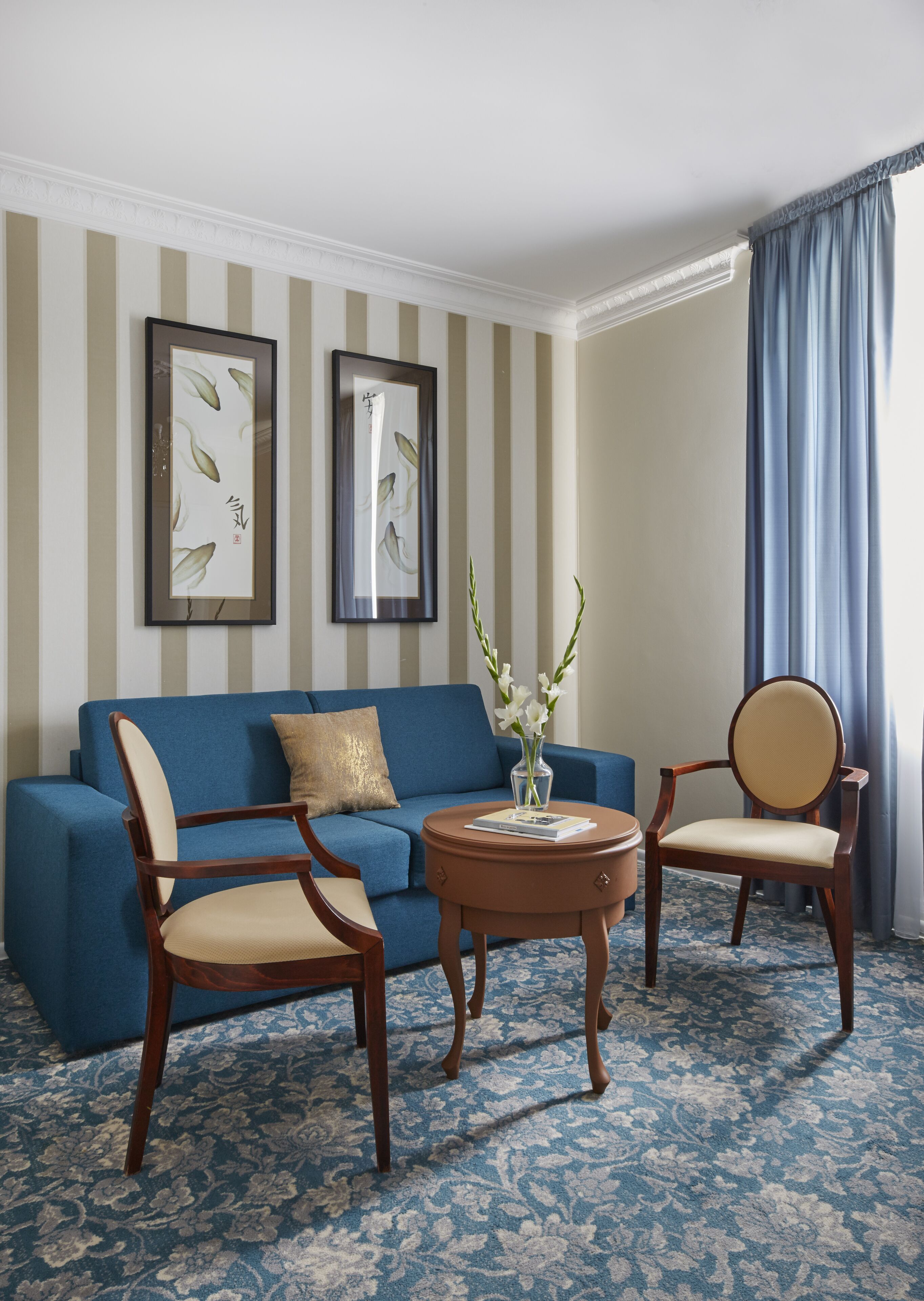 A room with a blue sofa, two wooden chairs, a round coffee table, and two framed artworks on the wall.