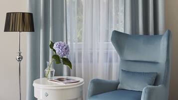Una habitación con un sillón azul, una mesita de noche blanca con un florero y un libro, y cortinas translúcidas sobre una ventana.