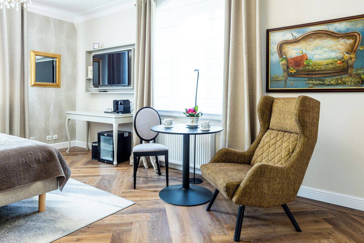 A hotel room with a bed, a desk with a computer, a round table with a vase, a chair, and a painting on the wall.
