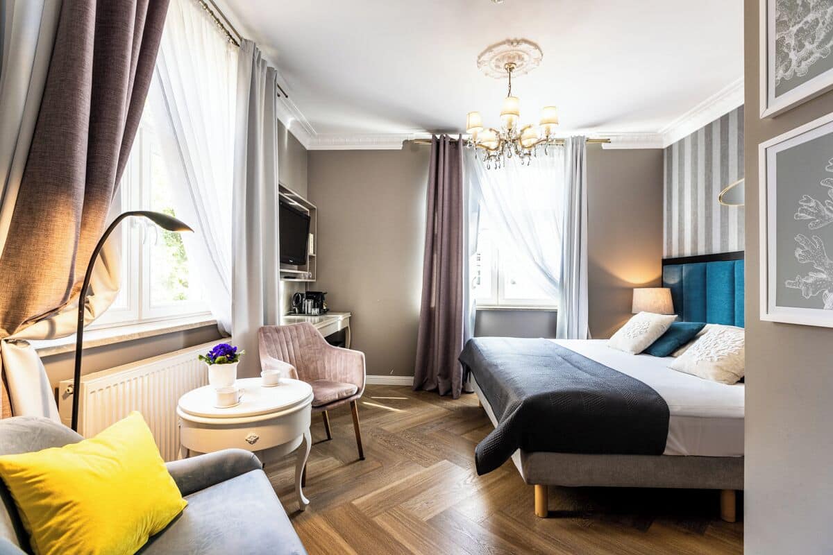 A hotel room with a bed, a desk, a chair, a small table, and a radiator.