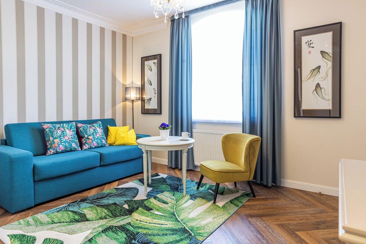 A living room with a blue sofa, yellow cushions, a round white table, a yellow armchair, a patterned rug, and two framed artworks on the walls.